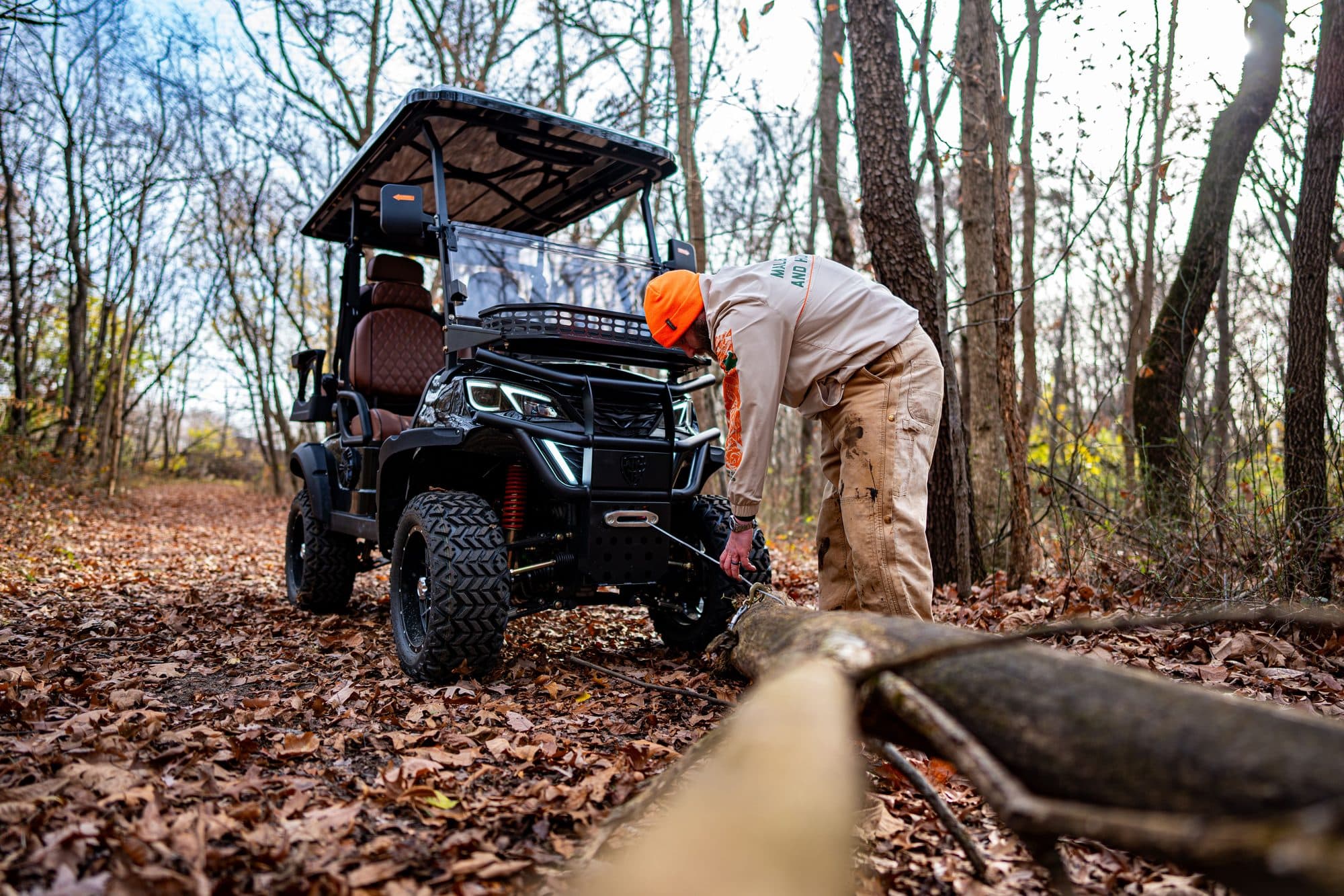 Black EA4X4 72V four-seat four-wheel-drive lithium electric golf cart using its front winch to pull a log in autumn woods, one adult in a blaze orange cap